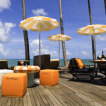 Neste imagem, a vista deslumbrante do mar e coqueiros como plano de fundo cria, um ambiente de luxo e elegância, exaltado pelas cores marca da Veuve Clicquot que orna o com bancos, guarda-sois, almofadas, garrafas e taças.
