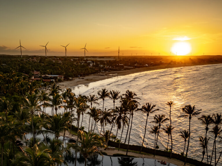 Pôr do sol dourado refletindo no mar tranquilo, com palmeiras contornando a paisagem e uma piscina à beira da praia. Ao fundo, aerogeradores se destacam no horizonte, compondo um cenário paradisíaco de natureza e sustentabilidade.