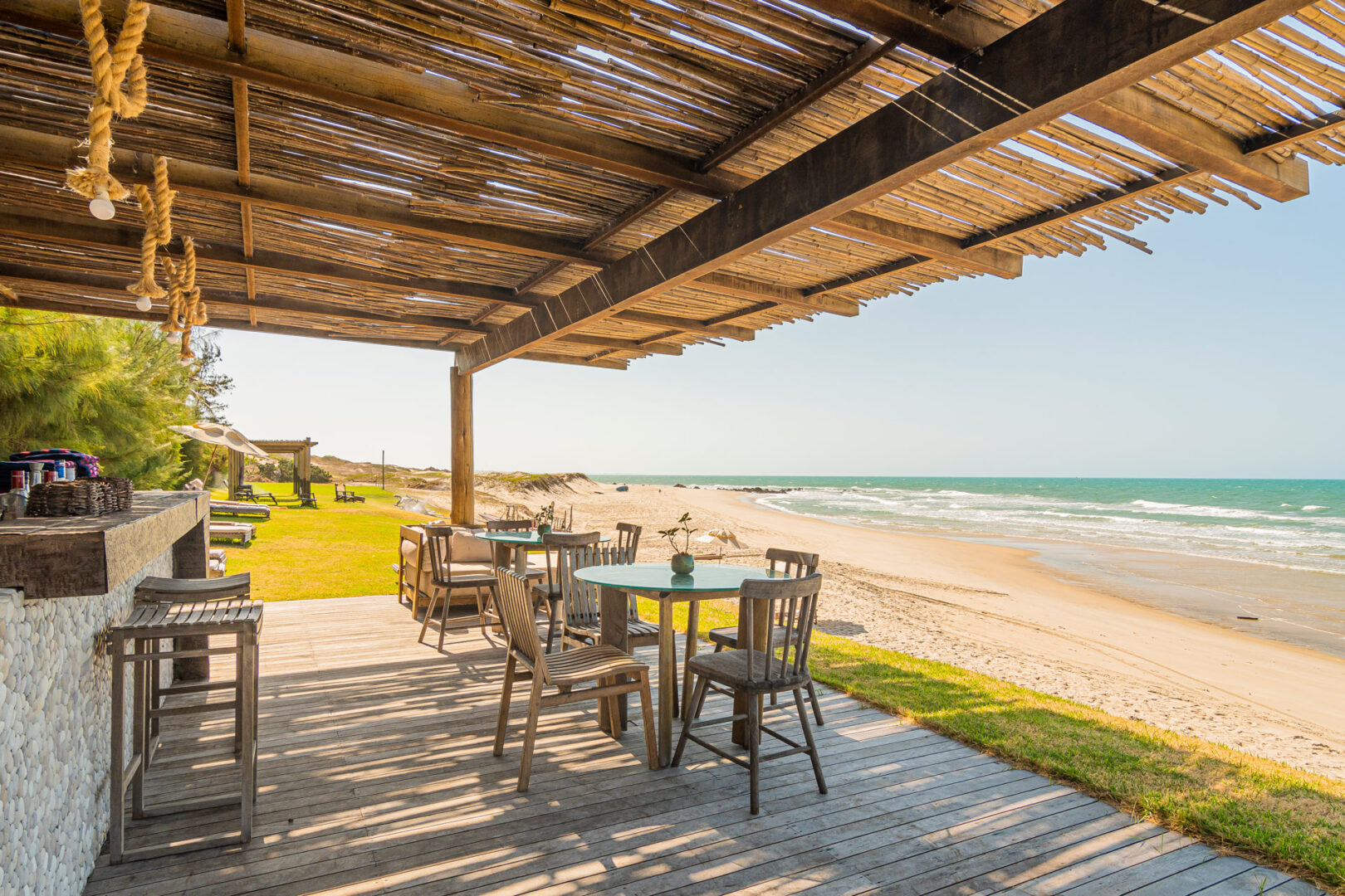 Bar de praia com estrutura de madeira e palha, rodeado por coqueiros. Mesas e cadeiras ficam dispostas na areia, de frente para o mar em uma das melhores praias do Ceará.