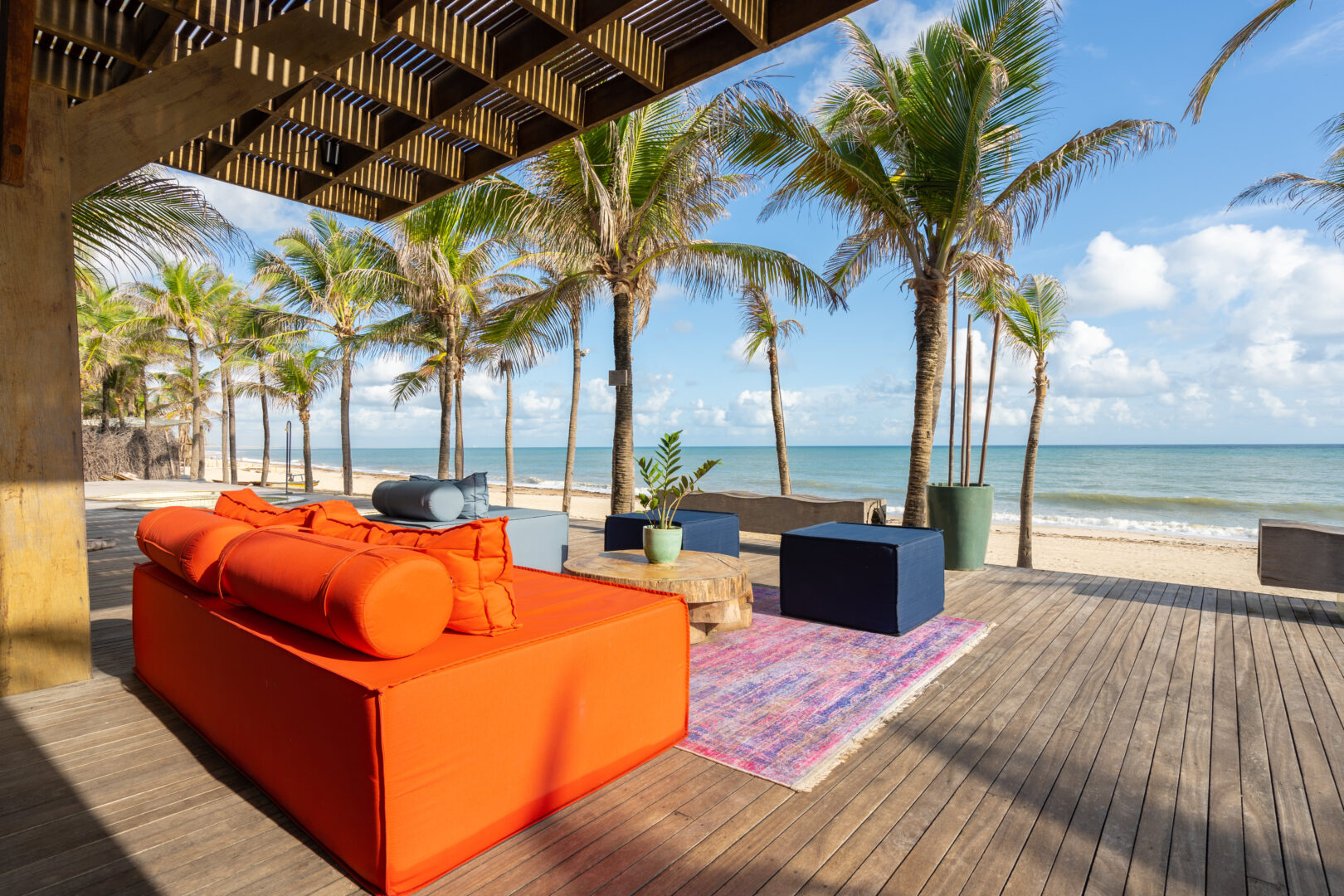 Espaço de lounge à beira-mar com deck de madeira, sofá laranja vibrante, pufes azuis e mesa de centro em madeira natural sobre um tapete colorido de frente para uma das melhores praias do litoral. O ambiente é sombreado por uma cobertura ripada que projeta sombras geométricas no chão. Ao fundo, palmeiras, areia clara e o mar calmo sob um céu azul com algumas nuvens, criando um cenário descontraído, sofisticado e convidativo para momentos de relaxamento à beira da praia.