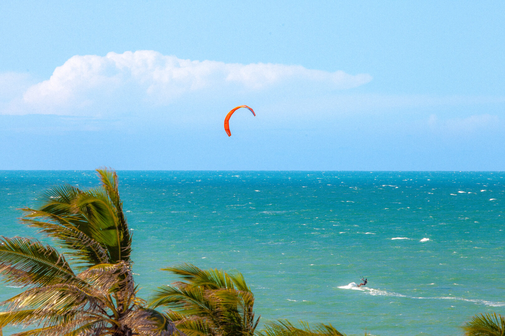 Prática de kitesurf na Praia do Cumbuco. Kitesurfista veleja pelo mar azul e esverdeado.