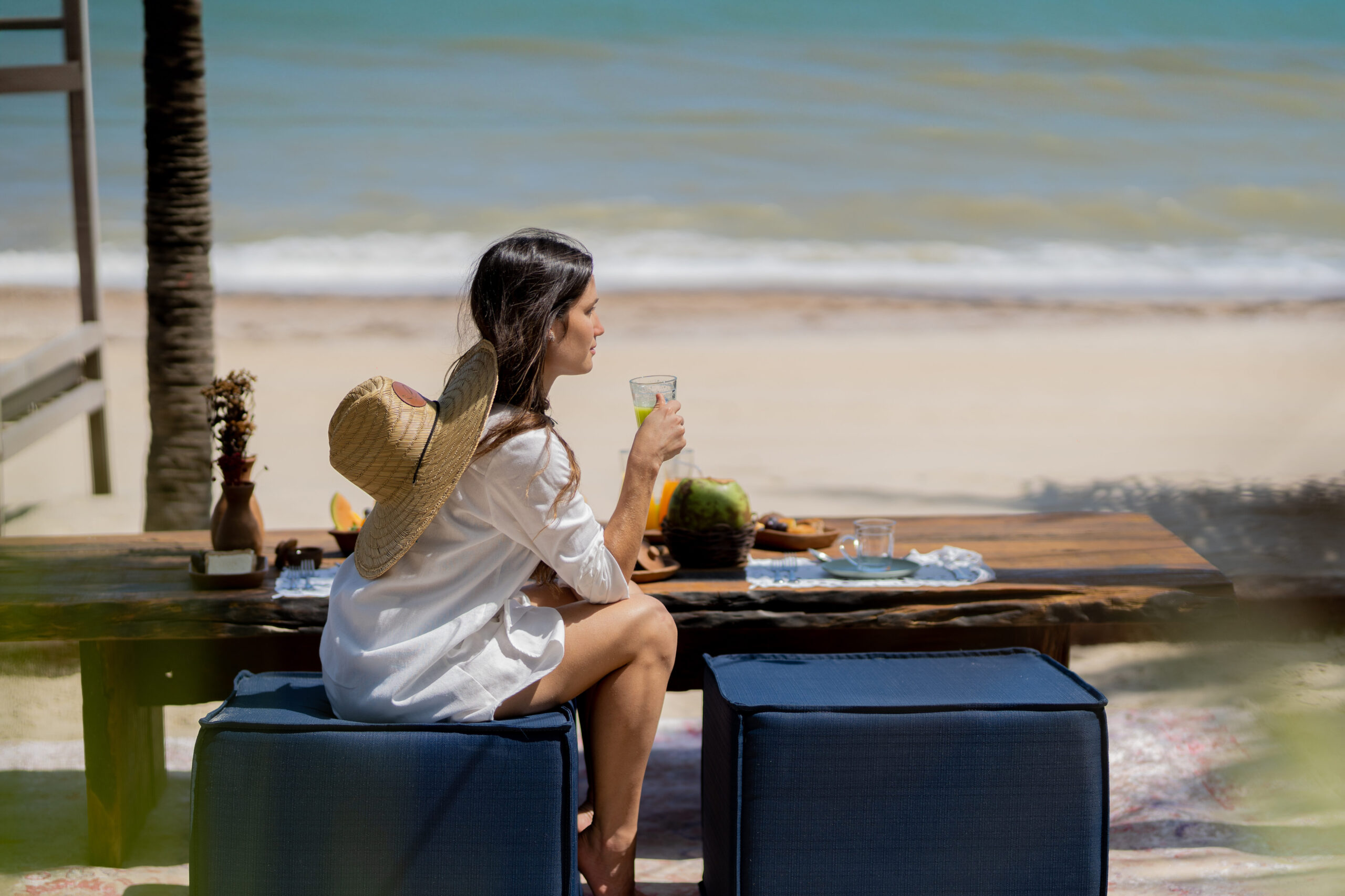 Mulher sentada em uma poltrona na areia da praia enquanto toma um café da manhã de frente para o mar e com os pés na areia