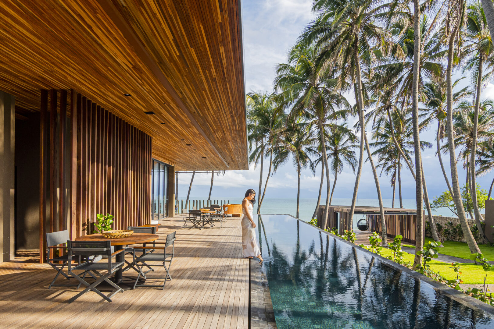 Mulher tocando pé na água da piscina sonorizada de um resort de luxo com a paisagem de coqueiros ao lado, um resort restaurante sofisticado com acabamento de madeira do outro lado colado a piscina e o mar ao fundo
