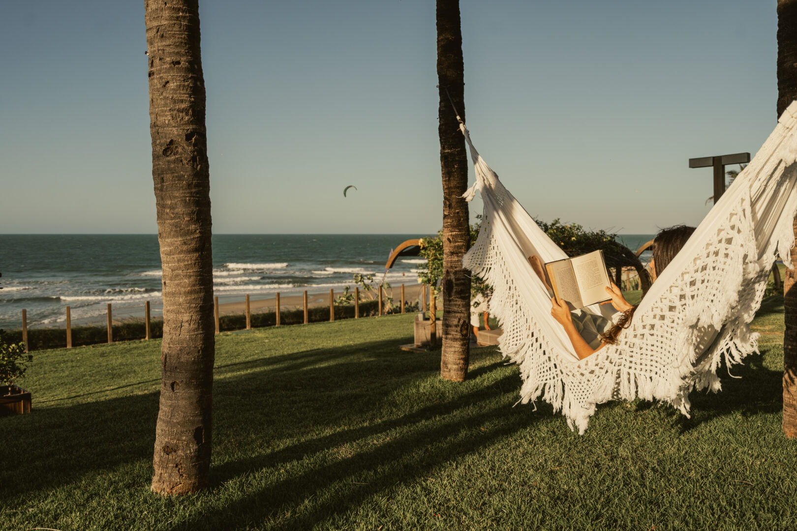 Mulher deitada em uma rede branca armada entre dois coqueiros aproveitando a brisa do mar enquanto lê um livro em sua viagem solo