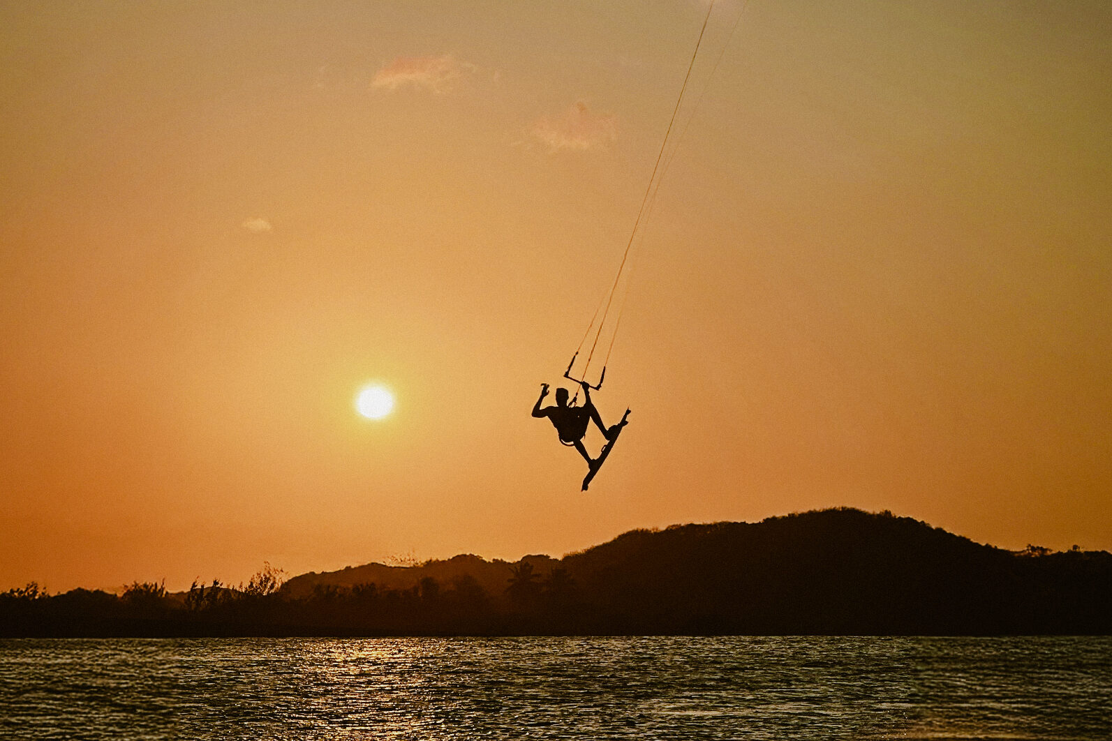 Ceará em janeiro: praticante de kitesurf salta sobre a água ao pôr do sol, com dunas ao fundo e céu alaranjado