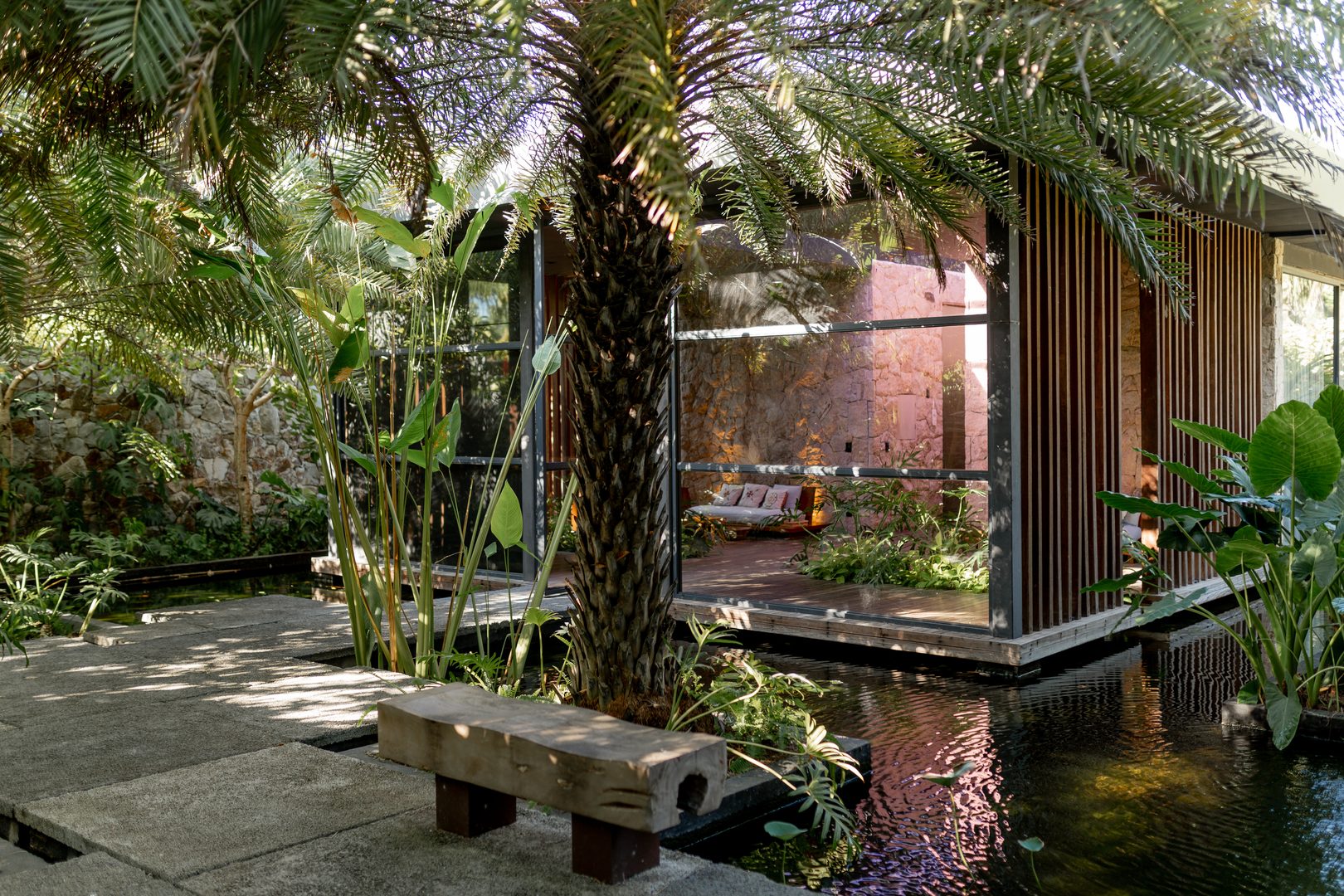 Fachada do spa com paredes de vidro e detalhes em madeira, cercada por vegetação tropical e espelho d’água.