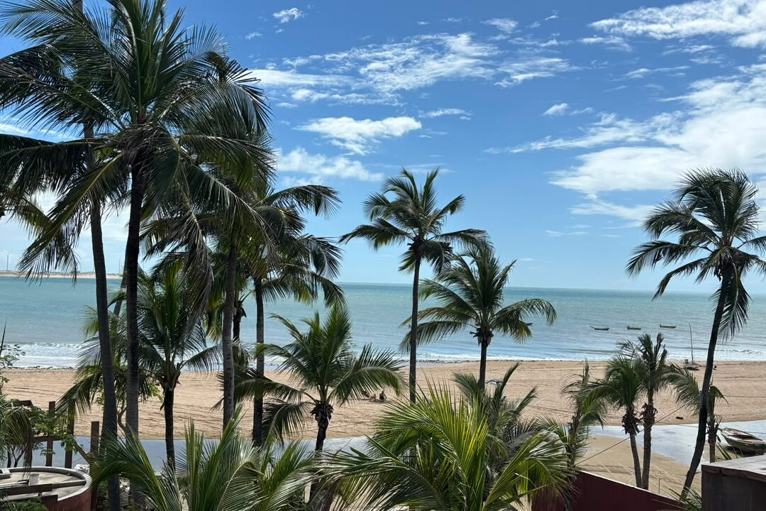 Ceará em janeiro: praia com coqueiros, faixa de areia e pequenas embarcações no mar sob céu azul com nuvens.