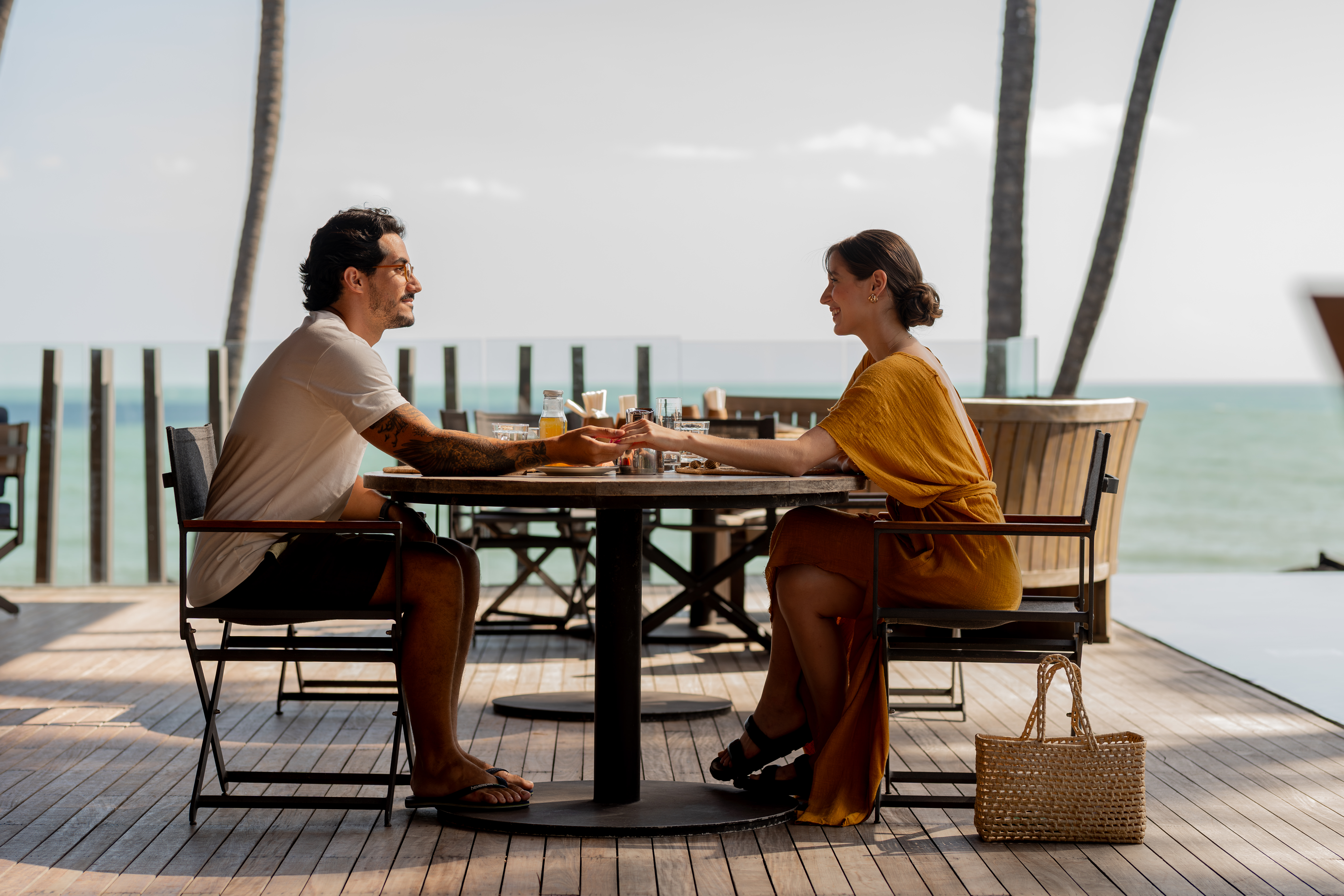 Ceará em janeiro: casal de mãos dadas à mesa em deck de madeira, com café da manhã servido e vista para o mar.