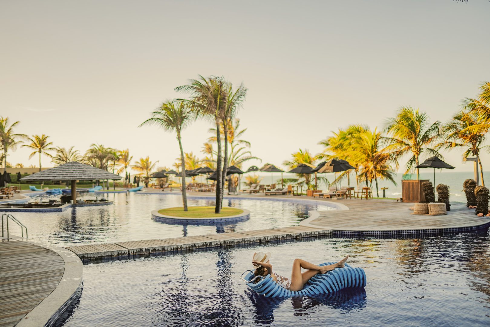 Ceará em janeiro: mulher relaxa em espreguiçadeira flutuante na piscina, cercada por coqueiros, com o mar ao fundo.